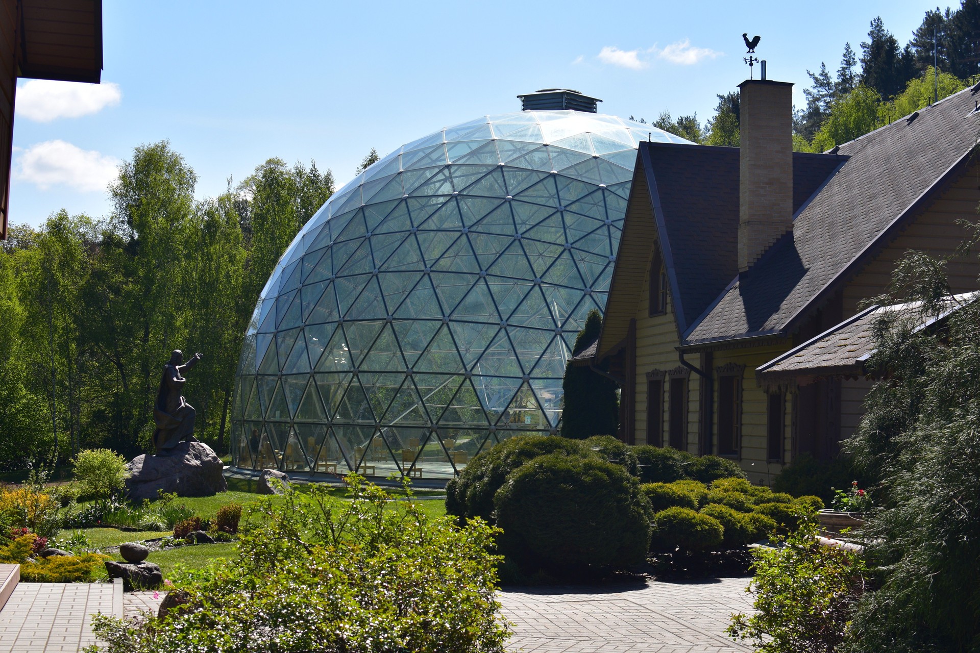 Glass geodesic dome surrounded by nature, next to a traditional wooden house. A statue stands in the garden, blending modern architecture with natural and cultural elements.